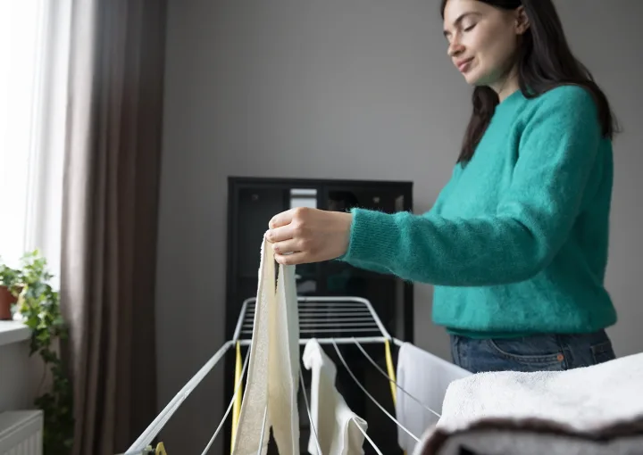 mulher estende roupa num estendal dentro de casa junto à janela e aquecedor